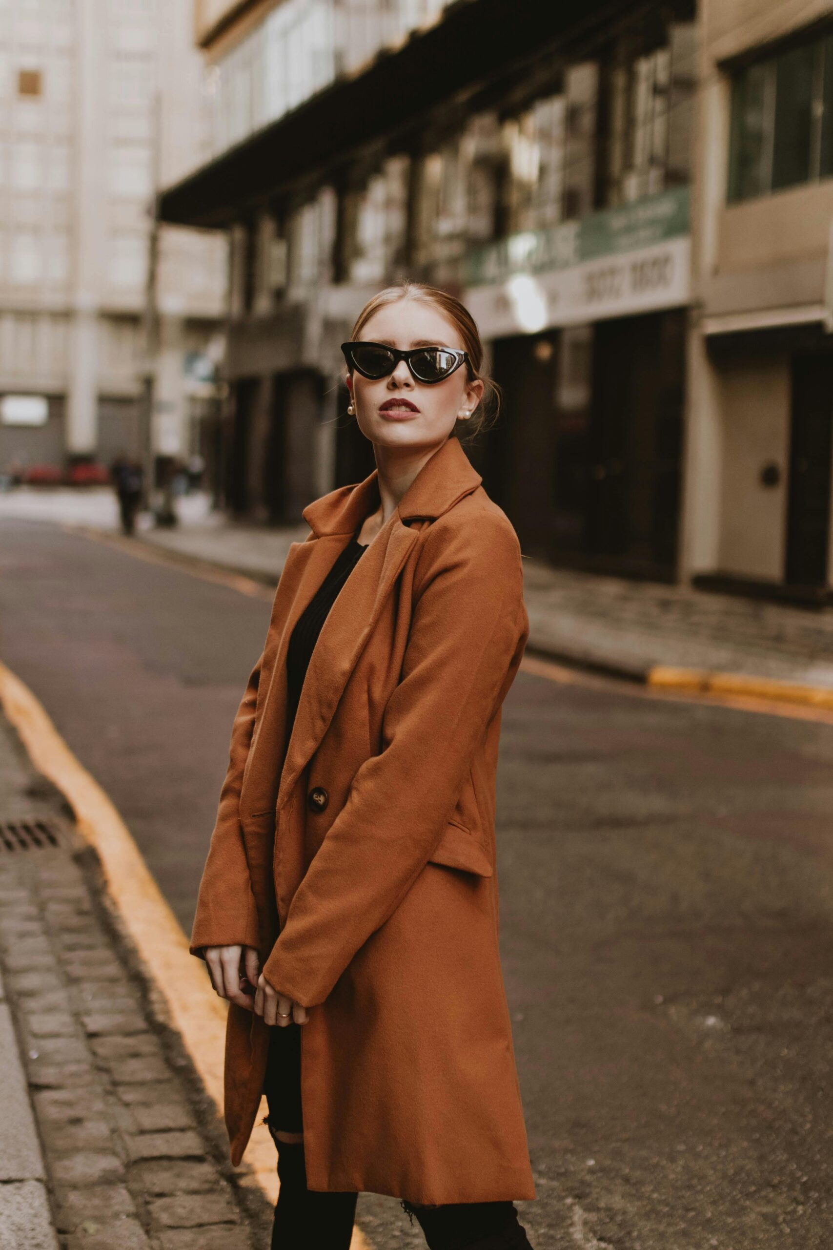 A woman standing in the street wearing brown coat and sunglasses.