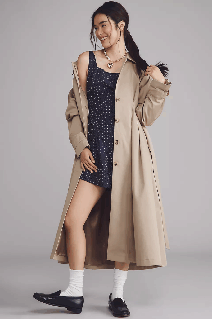 Model smiling wearing brown trench coat and blue sundress. 