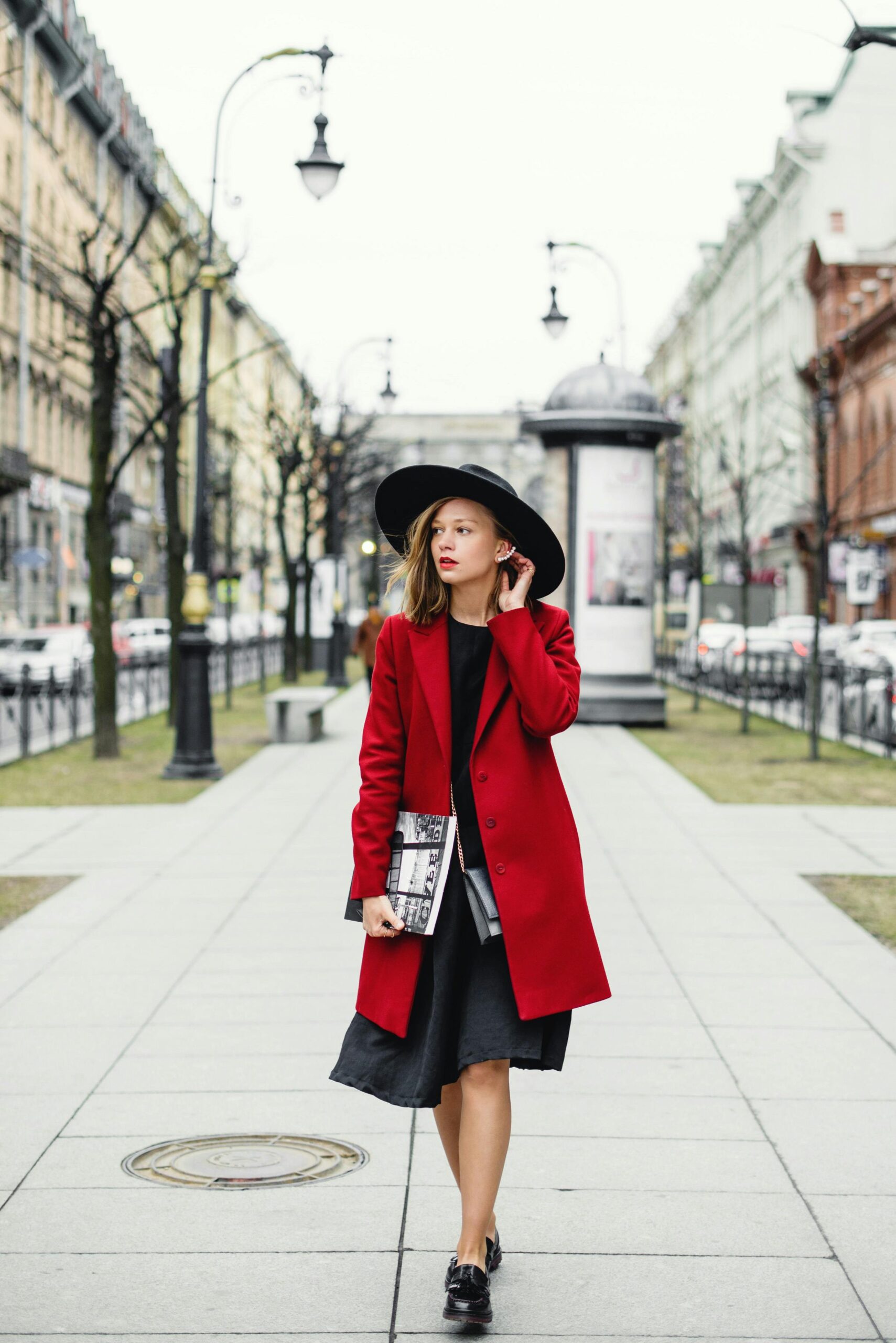 A woman wearing a black dress and a red blazer stands in the middle of a road.