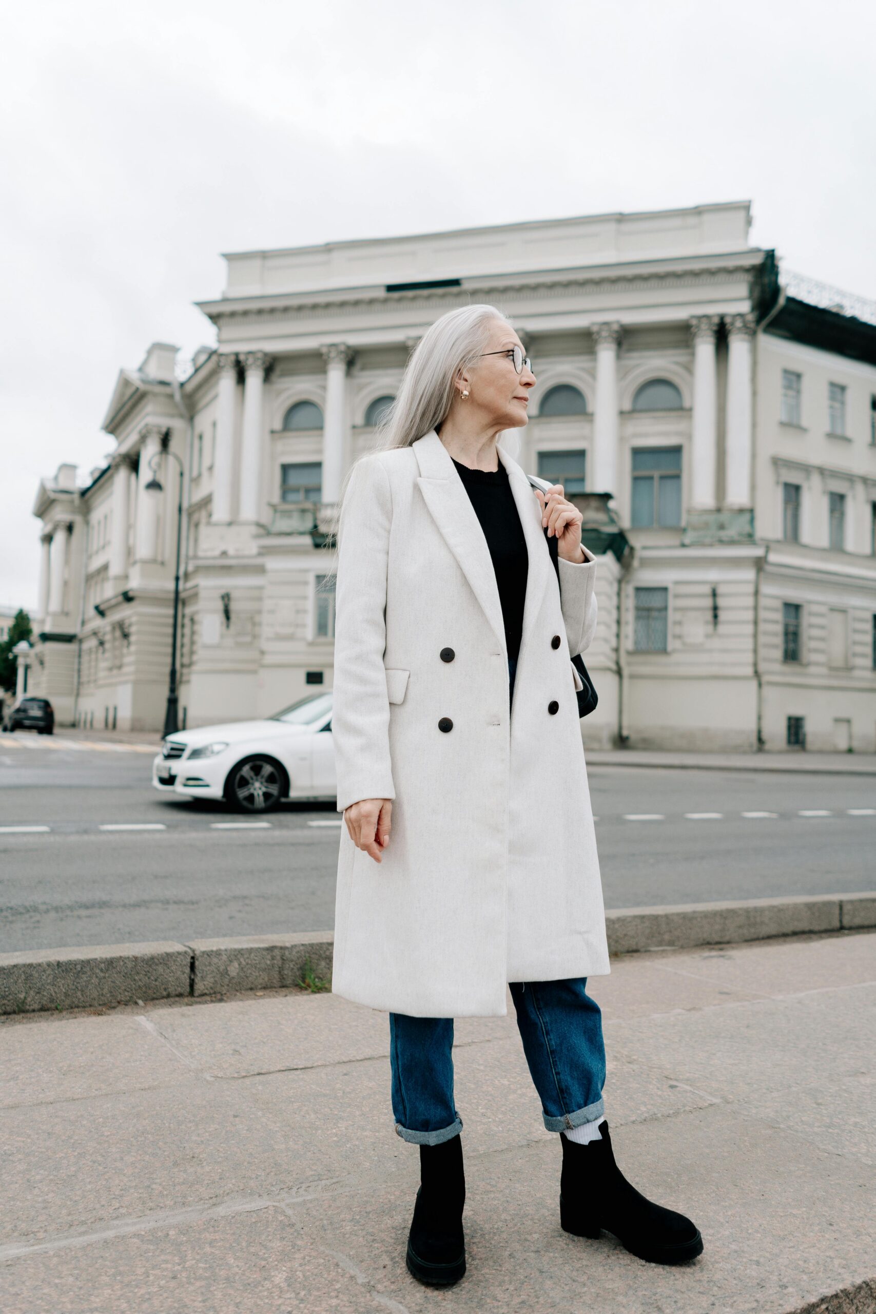 A woman standing in the street wearing a white coat, jeans, and boots.